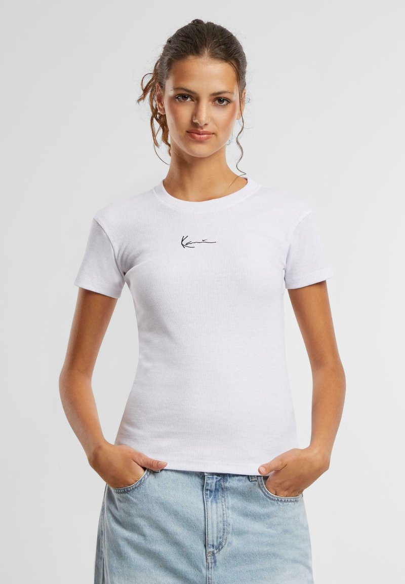 T-shirt blanche ajustée à manches courtes, texture côtelée, avec un logo noir signature sur la poitrine. Associé à un jean bleu clair.