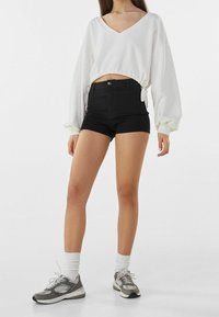 Vit croppad sweatshirt med V-hals och snördetalj, kombinerat med högmidjade svarta shorts och grå sportskor med vita strumpor.