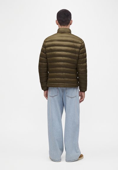 Homme portant une veste matelassée vert olive et un jean bleu clair à jambes larges, debout de dos sur un fond blanc uni.