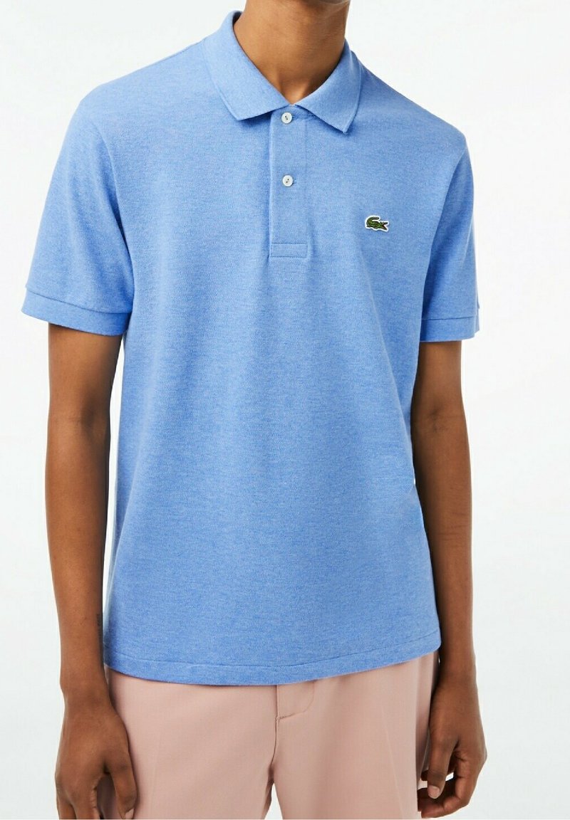 Personne portant un polo bleu clair à manches courtes avec un petit logo crocodile vert sur la poitrine gauche et un pantalon rose clair.