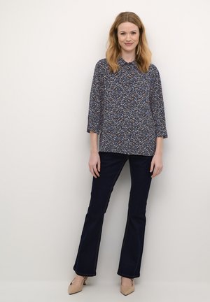Blusa floreale blu navy con maniche a 3/4, abbinata a jeans svasati scuri e tacchi beige. La blusa presenta un colletto con bottoni e un tessuto dalla trama particolare.
