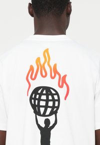 Silhueta de uma pessoa segurando um globo preto acima da cabeça, com chamas laranjas e vermelhas subindo atrás dele, em uma camisa branca nas costas.