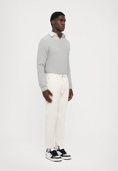 Suéter gris, camisa blanca de cuello debajo, jeans beige claros, zapatillas negras y blancas, de pie contra un fondo liso.