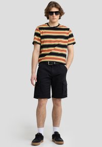 Stribet kortærmet t-shirt i sort, gul og rød; parret med sorte cargo shorts. Tilbehøret inkluderer sorte solbriller og sneakers.