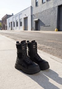 Black lace-up platform boots with thick soles placed on a city sidewalk near industrial buildings on a sunny day.