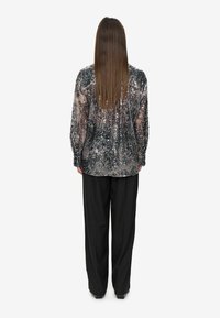 Femme avec de longs cheveux raides portant un haut à manches longues en sequins noirs et argentés et un pantalon noir ample, se tenant de dos.