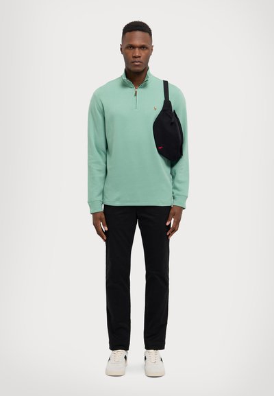 Pull vert clair à manches longues avec zip court, sac bandoulière noir, pantalon noir et baskets blanches avec accents noirs. Design simple.