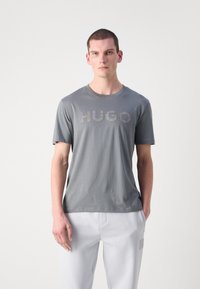 Pelēks īsām piedurknēm T-krekls ar teksturētu "HUGO" logotipu kontrastējošā printā. Audums šķiet mīksts ar brīvu piegriezumu.