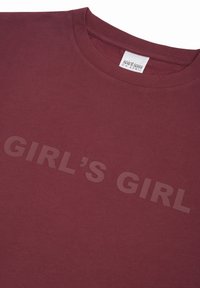 Tumši sarkana krekla ar apaļu kaklu, ar uzrakstu "GIRL'S GIRL" nedaudz tumšākā tumši sarkanā krāsā krūtīs un baltu zīmola etiķeti apkakles iekšpusē.