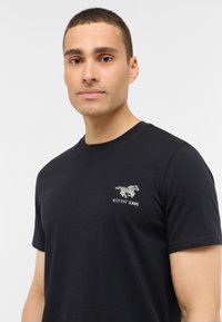 Czarna bawełniana koszulka z okrągłym dekoltem i krótkimi rękawami, ozdobiona szarym haftowanym logo Mustang Jeans na piersi.