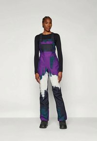 Lila und schwarze Latzhose mit Spritzern aus türkis und weißer Farbe. Verfügt über verstellbare Träger und wird mit einem schwarzen Langarmoberteil kombiniert.