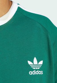 Green cotton t-shirt with a white ribbed collar and white Adidas logo featuring three leaves. Smooth texture and athletic fit design.
