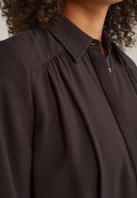 Chemise marron foncé avec un design plissé, dotée d'un col classique et d'une fermeture à boutons. Texture lisse avec un léger éclat et coupe cintrée.