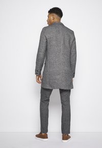 Manteau gris texturé à la coupe ajustée, avec poches latérales et une fente arrière simple, associé à un pantalon à carreaux assorti et des baskets marron.