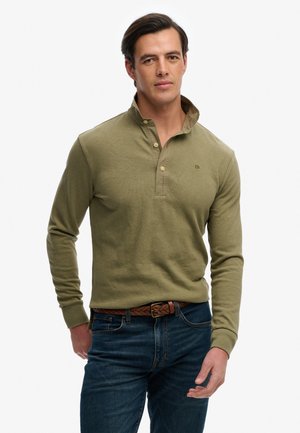 Mand iført olivengrøn langærmet polo med knapper, flettet brunt bælte og mørkeblå jeans, stående mod en hvid baggrund.