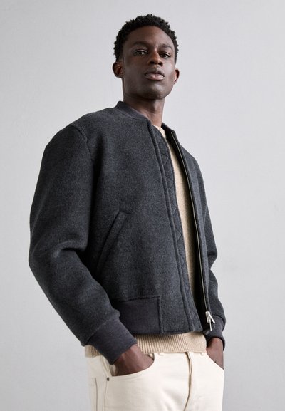 Chaqueta bomber gris oscuro con puños y dobladillo acanalados, con cierre de cremallera y bolsillos laterales, llevada sobre un suéter beige claro.