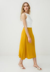 Gonna midi gialla in un tessuto leggero e fluido, abbinata a una canotta bianca aderente e sandali con tacco aperti bianchi. Design semplice.