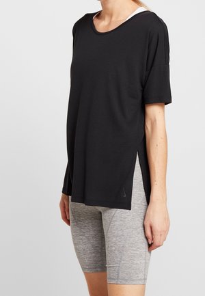 Frau trägt ein locker sitzendes schwarzes T-Shirt über einem weißen Sport-BH und eng anliegende hellgraue Sportshorts vor einfachem Hintergrund.