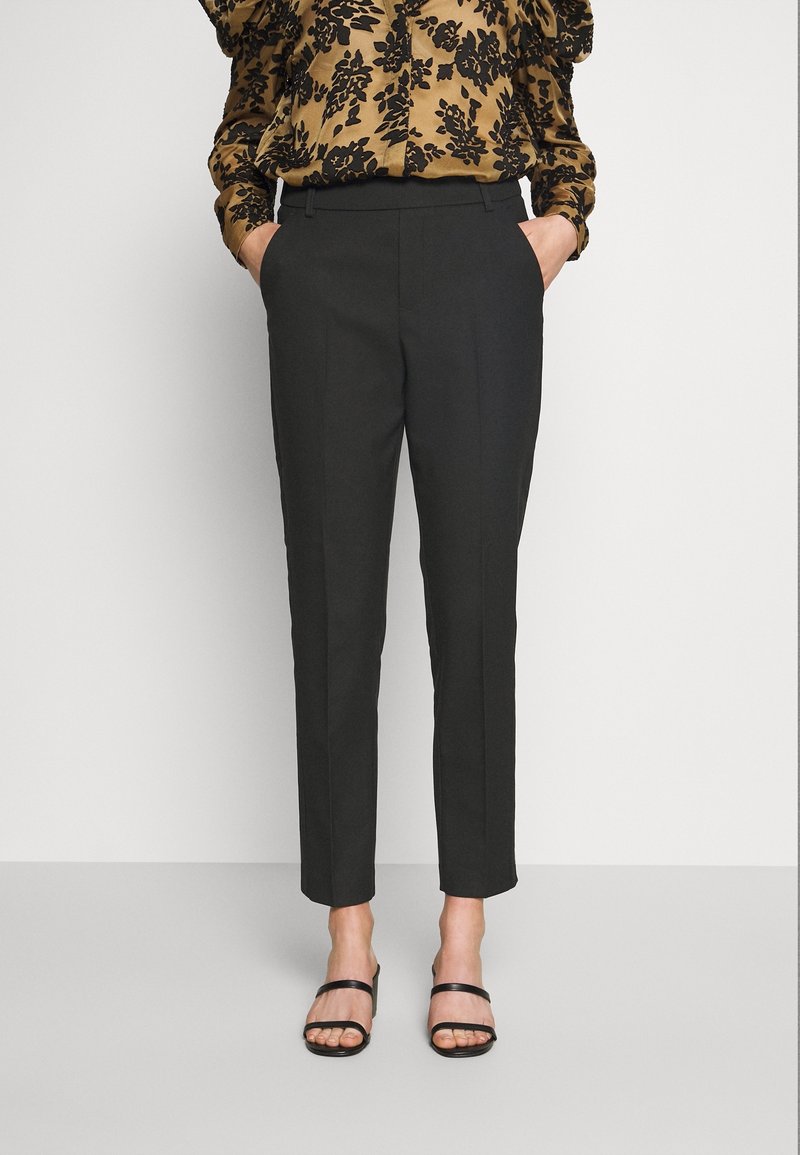 Pantalon noir sur mesure à coupe ajustée, fabriqué dans un tissu lisse. Comprend des poches latérales et un ourlet net. Porté avec un chemisier à motifs et des sandales.