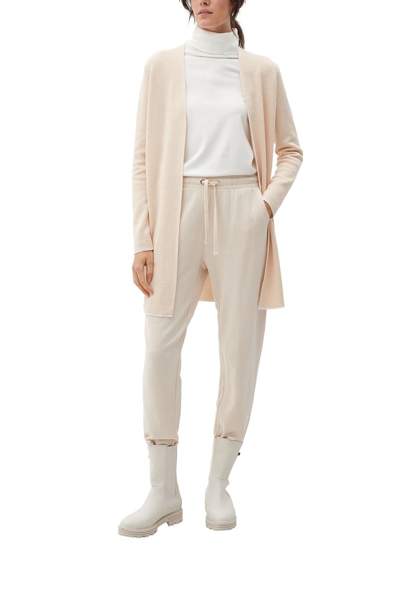 Femme portant un cardigan beige clair, un col roulé blanc, un pantalon beige à cordon et des bottines écrues, debout, une main dans la poche.