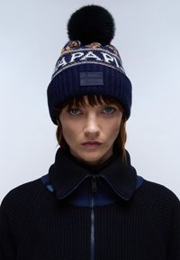 Berretto lavorato a maglia in blu navy con fascia a contrasto a motivo e pompon nero. Presenta una toppa con emblemma e una texture a coste. Indossato con una giacca nera con colletto.