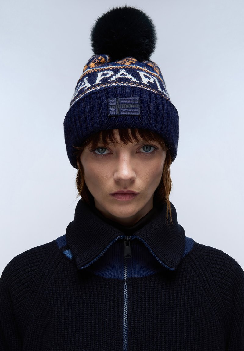 Berretto lavorato a maglia in blu navy con fascia a contrasto a motivo e pompon nero. Presenta una toppa con emblemma e una texture a coste. Indossato con una giacca nera con colletto.