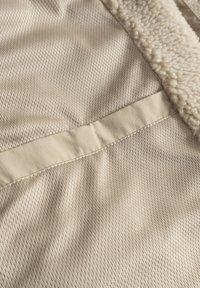 Beige stof med et struktureret netmønster, der har en glat kant og en fuzzy, lysfarvet foring langs kanten.