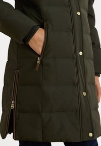 Cappotto isolato verde scuro con design trapuntato, dotato di tasche con zip, hardware color oro e polsini a costine per maggiore calore.
