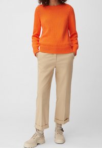 Donna che indossa un maglione a maglia arancione brillante, pantaloni beige con risvolto e stivaletti beige con lacci, in piedi con le mani nelle tasche.