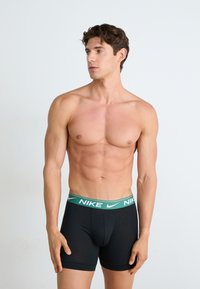 Schwarze Baumwoll-Boxershorts mit einem grünen Bund, der das Nike-Logo zeigt. Das Design ist enganliegend, hat eine weiche Textur und eine Länge bis zur Mitte des Oberschenkels.