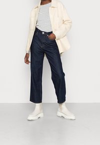 Hellbeige Cordjacke, gestreiftes Hemd, dunkle Jeans mit weitem Bein und weiße Ankle Boots mit strukturierten Sohlen, die einen Lagenlook kreieren.
