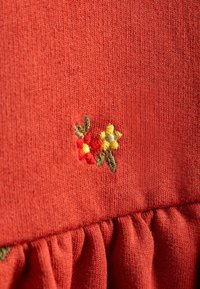 Červená pletená látka se shromážděným volánem na spodním okraji. Vyznačuje se barevným květinovým vyšíváním v červené, žluté a zelené barvě.