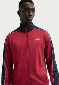 Κόκκινο αθλητικό μπουφάν Nike με σκούρες μπλε λεπτομέρειες, διαθέτει φερμουάρ μπροστά, ψηλό γιακά και λευκή επένδυση στα μανίκια. Ομαλή υφή υφάσματος.