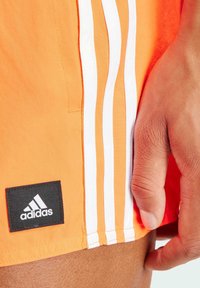 Pantalones cortos deportivos naranjas con tres rayas blancas a lo largo del costado y un parche del logo negro de Adidas en el dobladillo. Textura suave, material ligero.