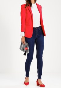 Blazer ajusté rouge sur un top blanc, associé à un jean skinny bleu foncé et des chaussures à talons blocs rouges. Tenant une pochette à motif zigzag noir et blanc.