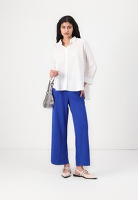 Chemise boutonnée blanche oversize, pantalon large bleu, chaussures nues à enfiler avec des brides, et un sac bandoulière argent.