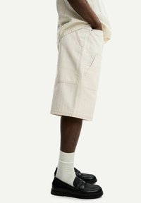 Shorts in denim beige con una finitura texture, con tasche frontali e un taglio dritto, abbinati a scarpe nere e calze bianche.