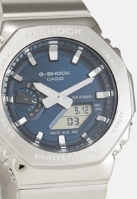 Montre G-SHOCK avec un cadran bleu, un boîtier en acier inoxydable argenté, un affichage numérique et plusieurs boutons de fonction. Résistante à l'eau jusqu'à 20 bars.