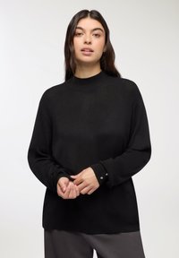 Maglione nero lavorato a maglia con collo alto, maniche lunghe e dettagli sottili ai polsini. Texture liscia, vestibilità rilassata. Indossato con pantaloni grigi.