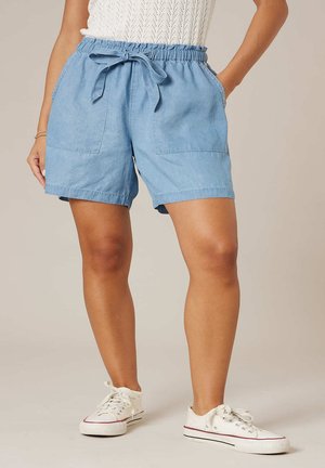 Femme portant un short à taille élastique bleu clair noué devant avec poches latérales et baskets blanches, debout avec une main dans la poche.