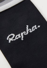 Calcetín de tela negra con un patrón de punto texturizado, con texto bordado en blanco que dice "Rapha". La parte superior tiene un acento a rayas en contraste.