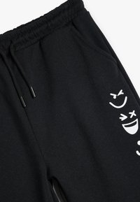 Sorte sweatpants lavet af blødt stof, med en snøre i taljen og sidelommer, og med hvide cartoon ansigt designs på venstre ben.