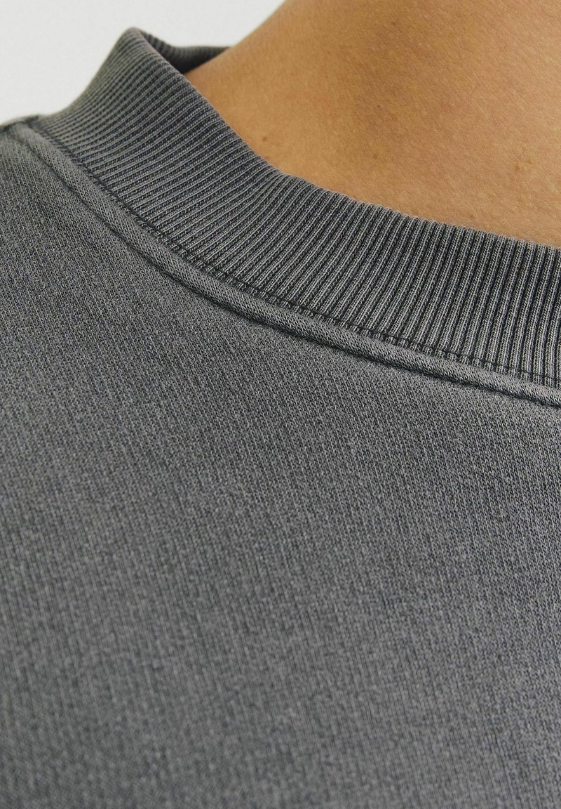 Šedá mikinová látka s texturovaným, žebrovaným límcem. Měkký, hladký povrch s mírným leskem, zašité švy jsou viditelné poblíž límce.