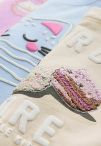Drie pastelgekleurde sweatshirts hebben speelse ontwerpen: een blauwe met een kat, een roze met een cupcake en een beige met een textuurmotief van een dessert.