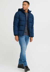 Marineblå pufferjakke med hette, kviltet design, to sidelommer, brukt over en grå genser, parret med blå jeans og svarte støvler.