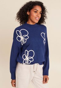 Pull en tricot marine avec des motifs floraux brodés blancs, col montant et manches longues, associé à un pantalon de couleur claire. Texture douce.