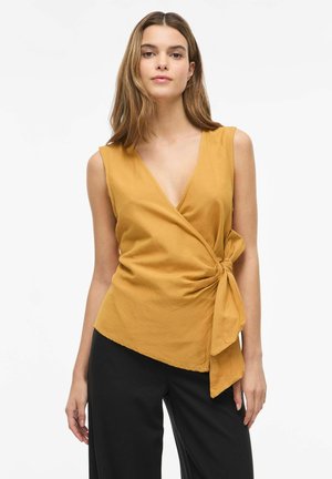 Femme portant un haut cache-cœur sans manches jaune moutarde avec un nœud sur le côté et un pantalon noir, debout devant un fond blanc.