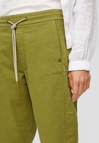 Pantalons vert olive avec une taille à cordon de serrage, dotés de poches latérales et d'un tissu lisse et léger. Des coutures discrètes sont visibles.