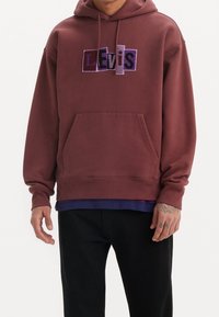 Osoba v bordovém mikině Levi's s multibarevným logem, černými kalhotami a viditelnými tetováními na rukou na bílém pozadí.