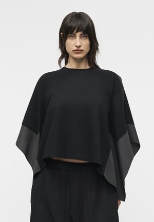 Γυναίκα με σκούρα μαλλιά που φοράει μια φαρδιά μαύρη μπλούζα με φαρδιά, διάφανα μανίκια και μαύρο παντελόνι, στέκεται μπροστά σε απλό λευκό φόντο.